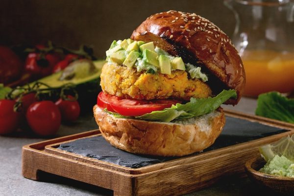 Vegan burger with carrot and avocado in classic bun, served on wooden slate board with avocado sauce and ingredients above on grey texture table. Homemade healthy fast food.